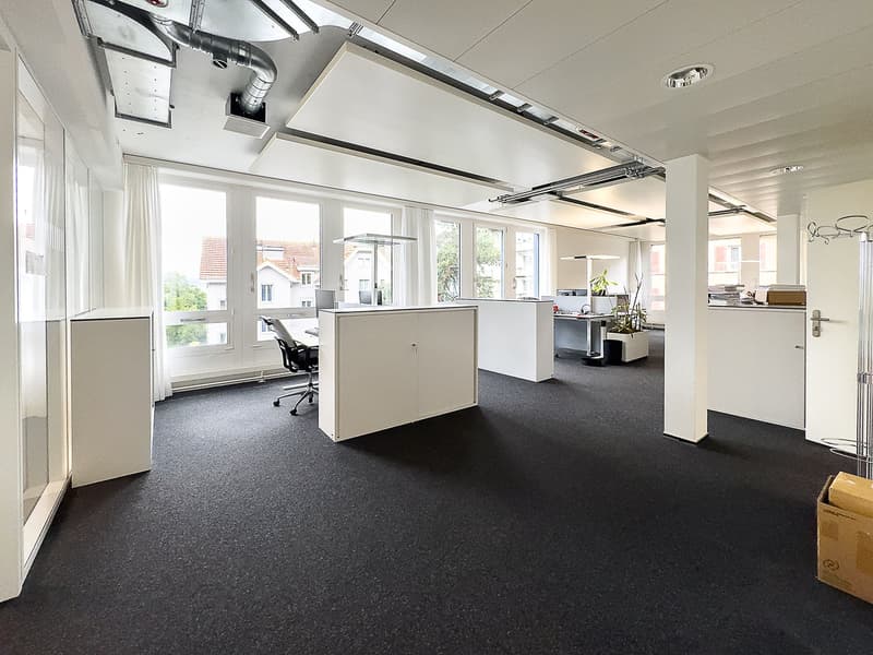 Open floor plan with a row of workstations facing windows, each with a desk, chair, and monitor, large windows allowing for natural light, modern furnishings with white walls and flooring, a box in the right side.