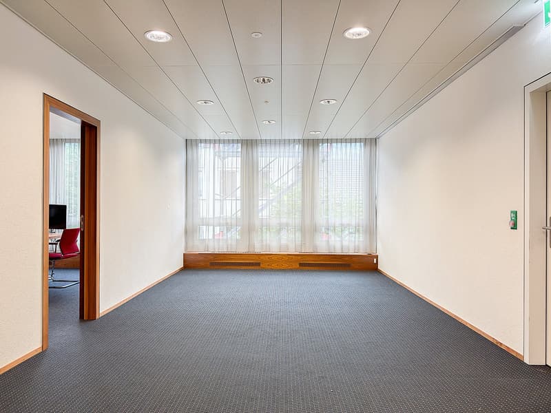 Empty room, white walls, carpeted floor, wooden window sill, large windows with white curtains, door to another room with red chair and monitor