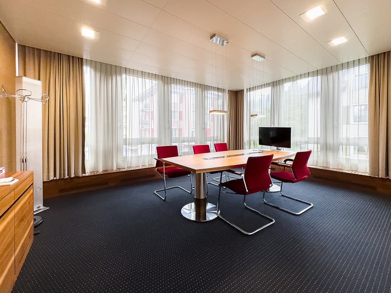 Empty room with a large table, 6 chairs, monitor, 3 large windows, wooden desk, wooden floor, white walls, ceiling lights