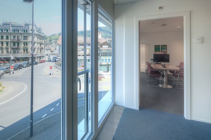 Large windows with a view of a city street, modern office space with a conference table and chairs, hardwood floors, and a TV mounted on the wall