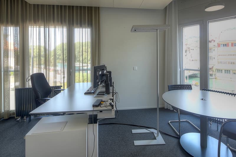 The image shows an office space with a large desk, computer setup, and chairs. The room has large windows with curtains, providing a view of the outdoors. The floor is carpeted, and the walls are painted a light color. The office appears to be well-equipp