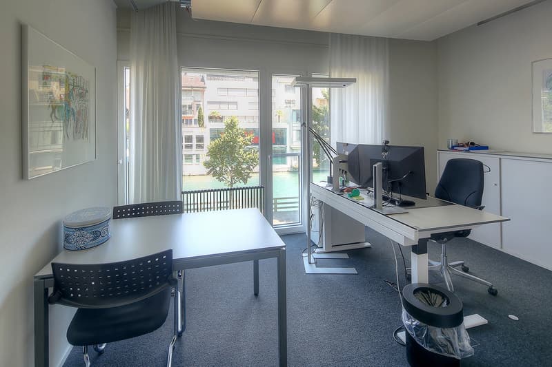 Spacious office with large windows overlooking a canal, modern furniture including a desk, chairs, and computer equipment, artwork on the walls