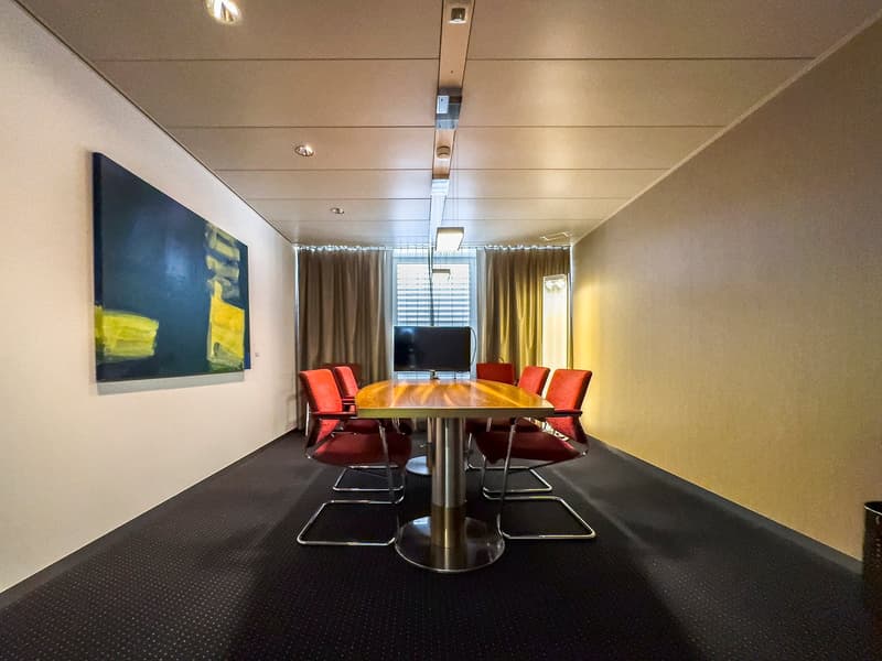 A conference room with a round wooden table, red leather chairs, a large painting on the wall, a television mounted on the wall, curtains covering a window, and an oval metal floor lamp