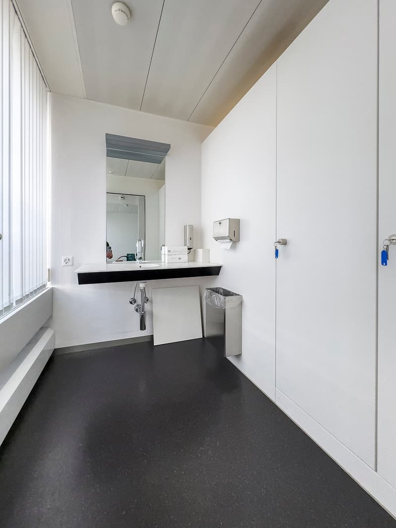 Modern, minimalist bathroom with white walls, black floor, a sink with faucet, a mirror, a trash can, and paper towel and hand soap dispensers
