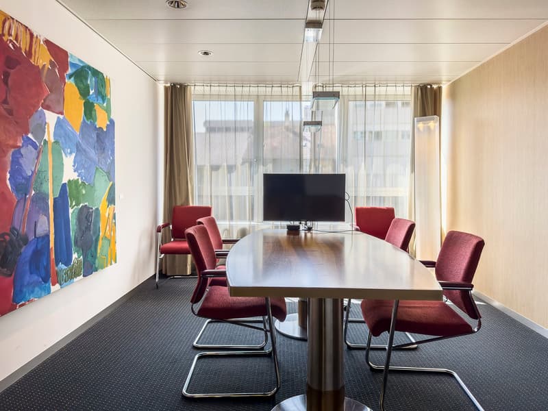 conference room with a long oval table, 6 red chairs, large windows with curtains, large abstract painting, monitor on the table