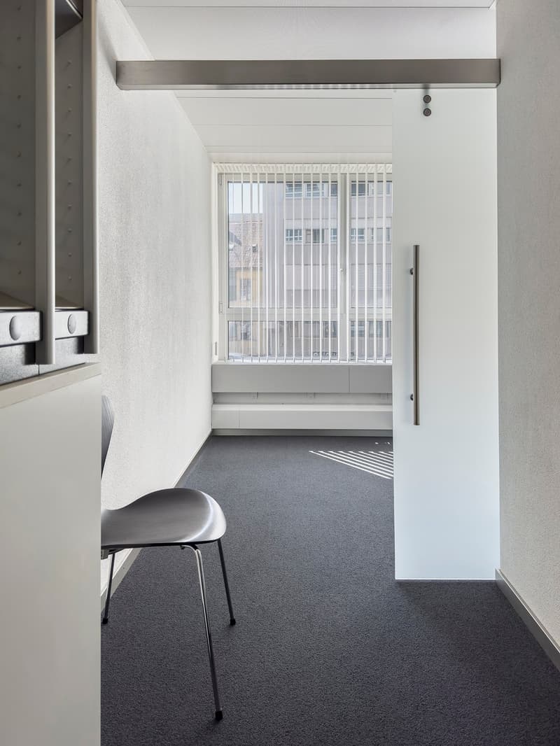 small empty room, white walls, gray floor, sliding door, large window with blinds, chair in corner