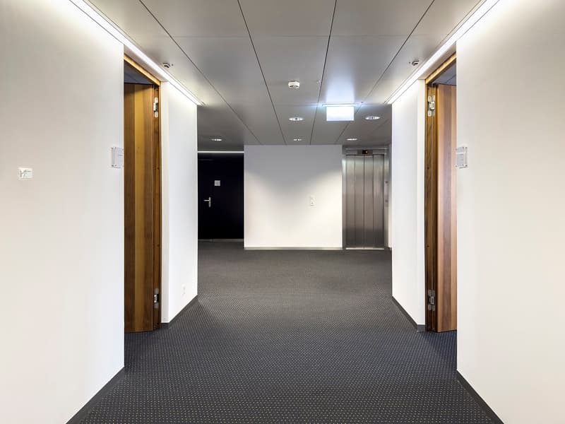 Empty white corridor with a carpeted floor, silver elevator in the middle, and two closed doors at the ends.