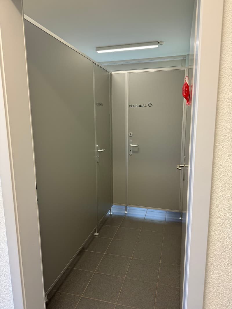 The image shows a hallway with two doors, one labeled 'Restroom' and the other labeled 'Personal'. The floor is tiled, and the walls are a light gray color. The lighting appears to be provided by a fluorescent light fixture.