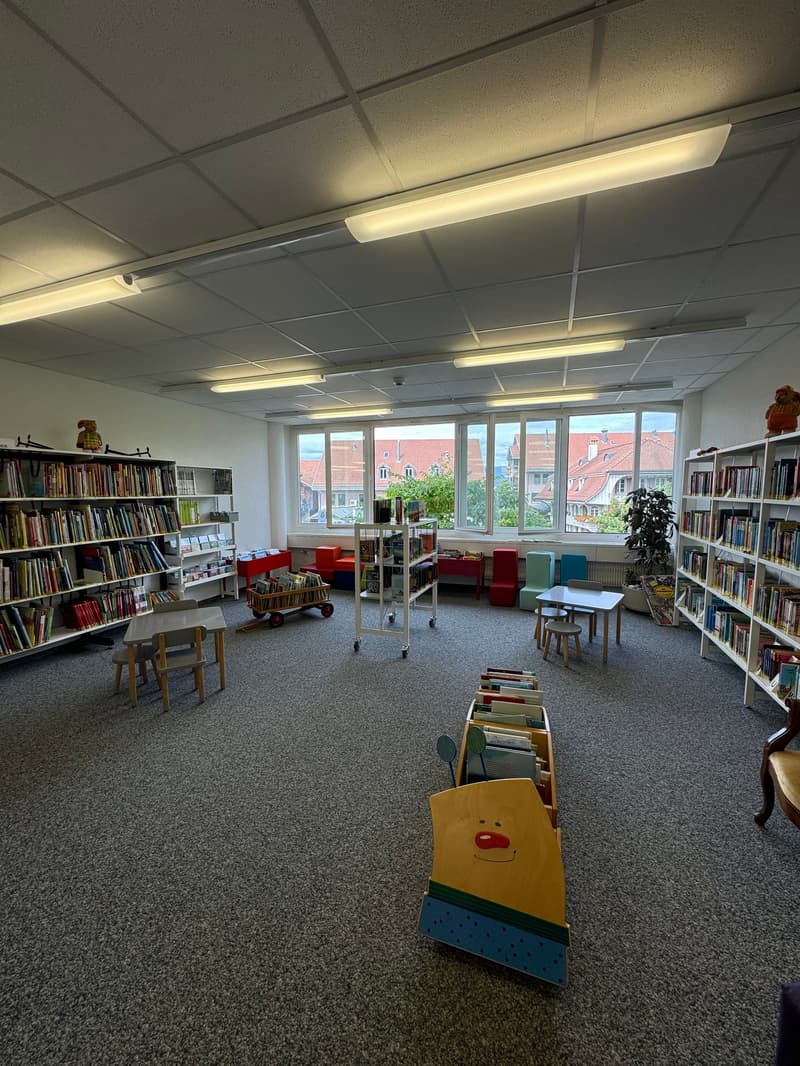 The image shows a library or study area with bookshelves lining the walls, various seating options including chairs and stools, and a large window overlooking the exterior. The room has a warm, inviting atmosphere with the lighting and decor.