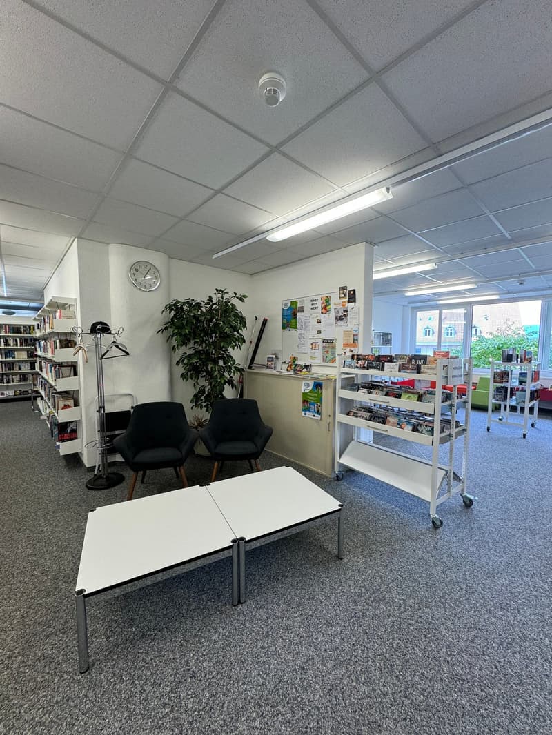 The image shows an office or study area with a table, chairs, bookshelves, and a plant. The room has a clock on the wall, and there are various items and materials displayed on the shelves. The space appears to be well-lit with overhead lighting and natur