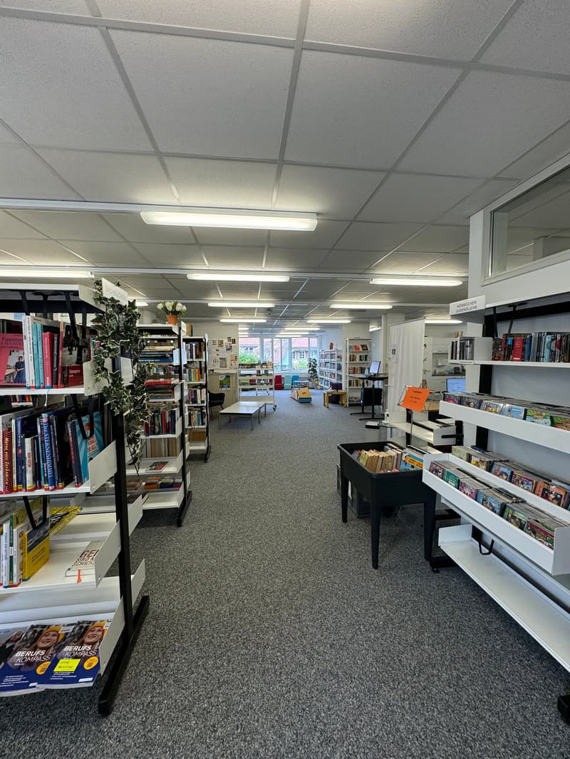 The image shows the interior of a bookstore. The space is filled with bookshelves and displays, with a variety of books and other merchandise available. The lighting is provided by fluorescent ceiling fixtures, and the floor is covered in a gray carpet. T