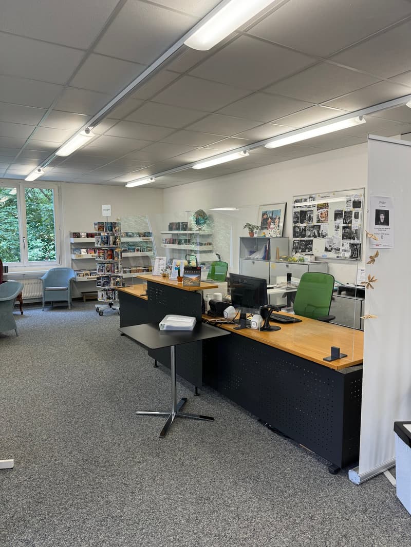 The image shows an office space with a reception desk, computer workstations, shelves with books and other items, and a bulletin board with posters and artwork. The room has a modern, professional appearance with gray carpeting, white walls, and overhead