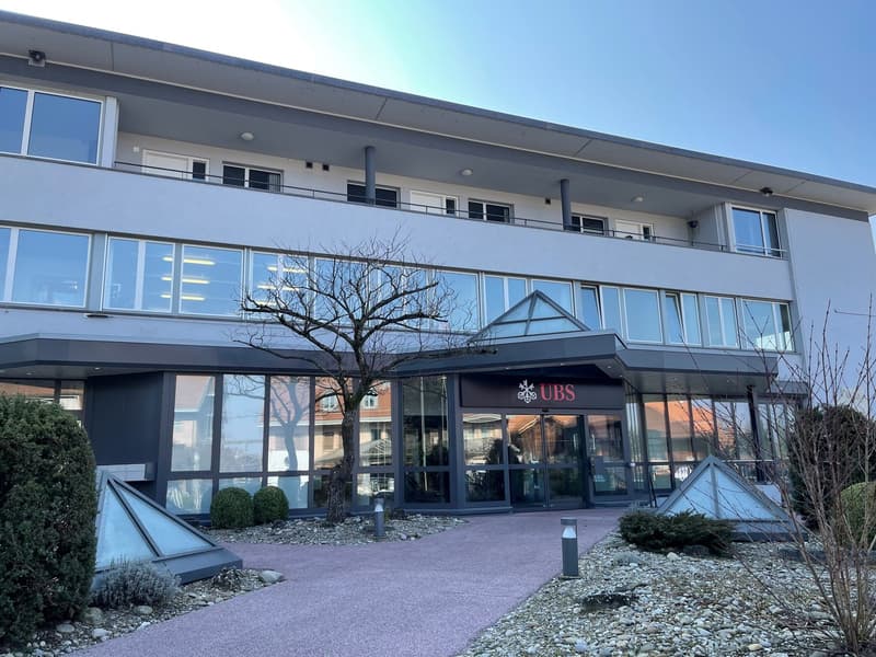 2-story commercial building with large glass windows, UBS bank signage, and a paved parking area in front