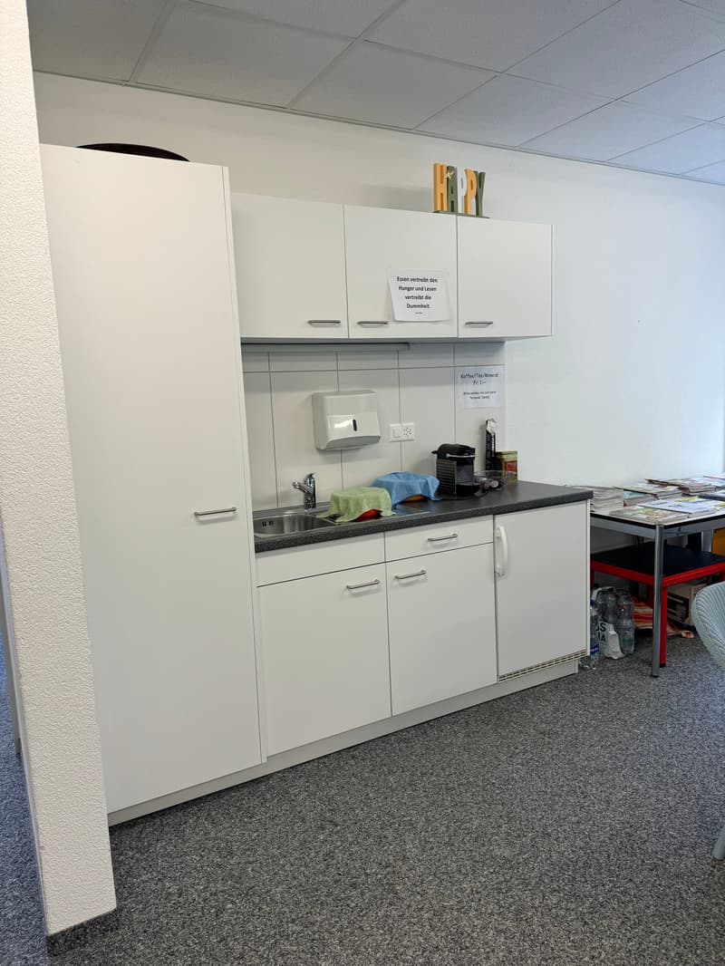 The image shows a small kitchenette area in an office or workplace setting. It has white cabinets, a sink, and a dishwasher. There are some cleaning supplies and a coffee maker visible on the counter.
