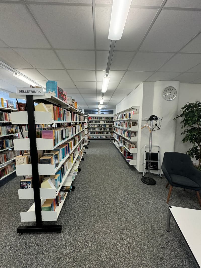 The image shows the interior of a library or bookstore. The space is filled with bookshelves lining the walls, creating long aisles for browsing. The shelves are stocked with a variety of books, and there is a clock and a chair visible in the space, sugge