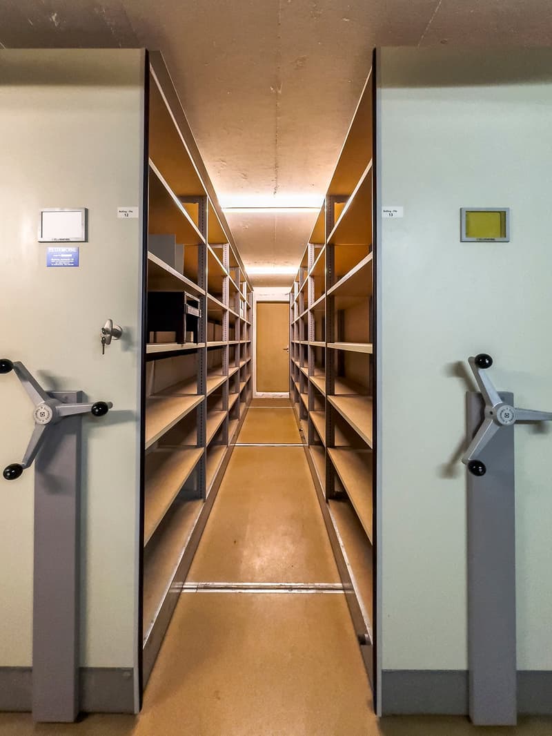 The image shows a long, narrow storage room with multiple shelves on both sides. The shelves appear to be made of metal and are used for storing various items. The room has a concrete floor and the walls are painted a light color. There are two metal cabi