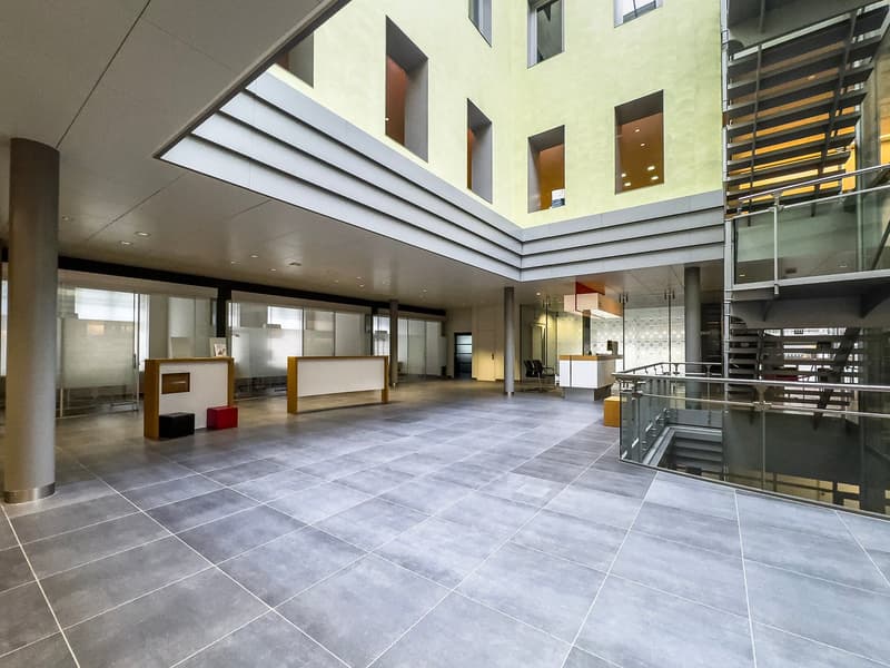 Large open lobby area with high ceilings, modern architectural design, gray tile flooring, glass walls, and built-in furniture elements