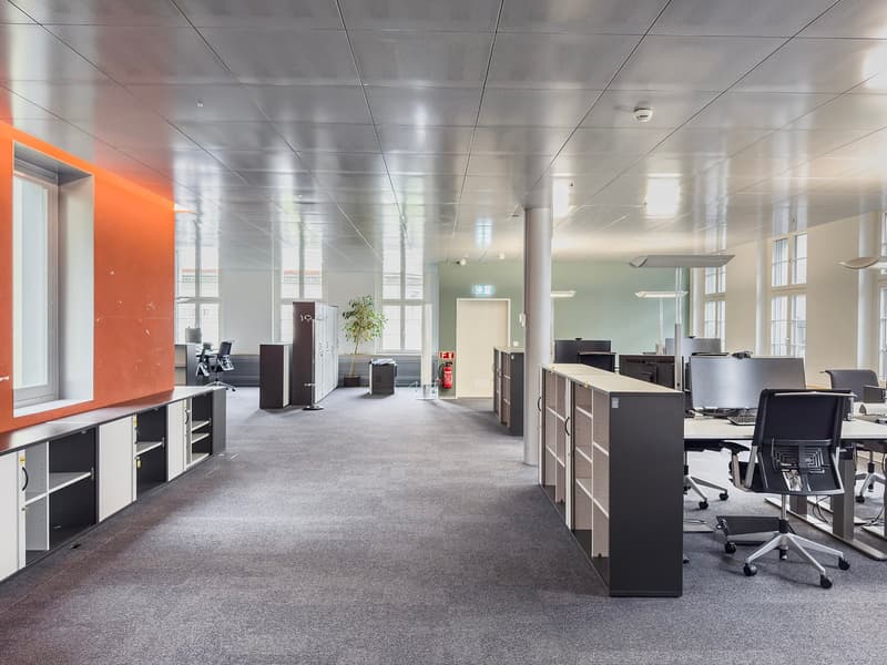 Large open office space with gray carpeted floor, white ceiling tiles, and black office furniture including desks, cabinets, and chairs. There is a red accent wall and some potted plants visible.