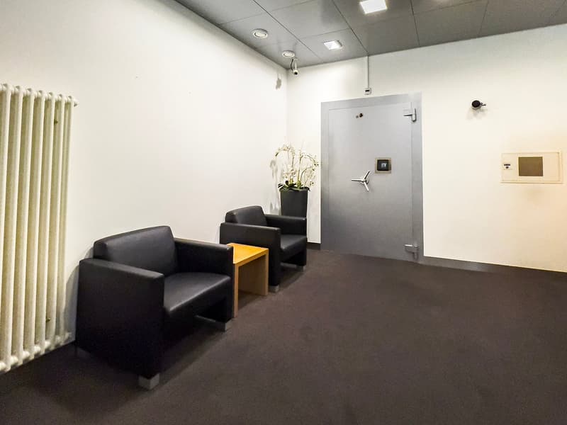 The image shows a small office space with a white wall, a gray ceiling, and black leather chairs arranged around a yellow side table. There is a security door and a security camera visible on the wall.