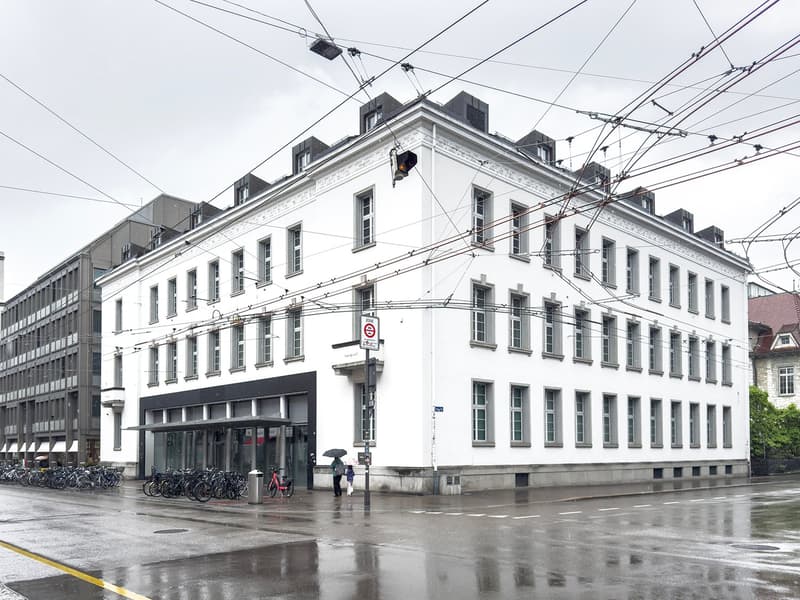 A multi-story white building with ornate architectural details, including windows and a prominent roofline. The building appears to be located in an urban area, with power lines and other infrastructure visible overhead. There are bicycles parked outside