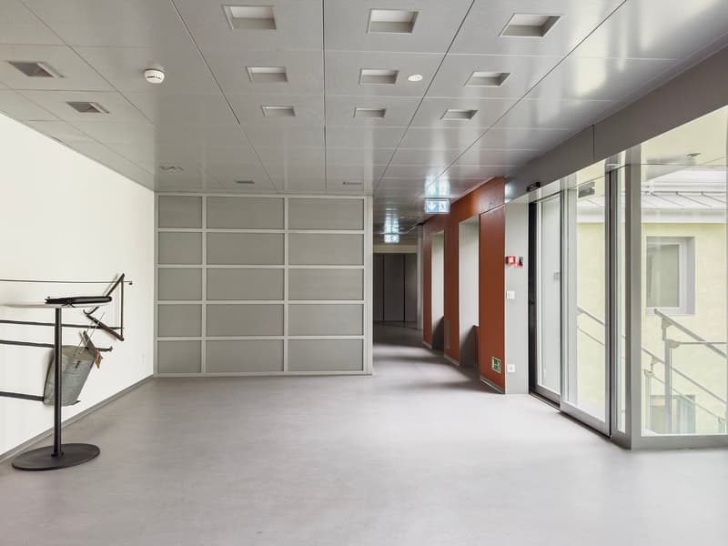 The image shows a modern office space with a long hallway. The walls are made of gray panels, and the ceiling has recessed lighting fixtures. There are glass doors and windows along the hallway, allowing natural light to enter the space. The floor is a li