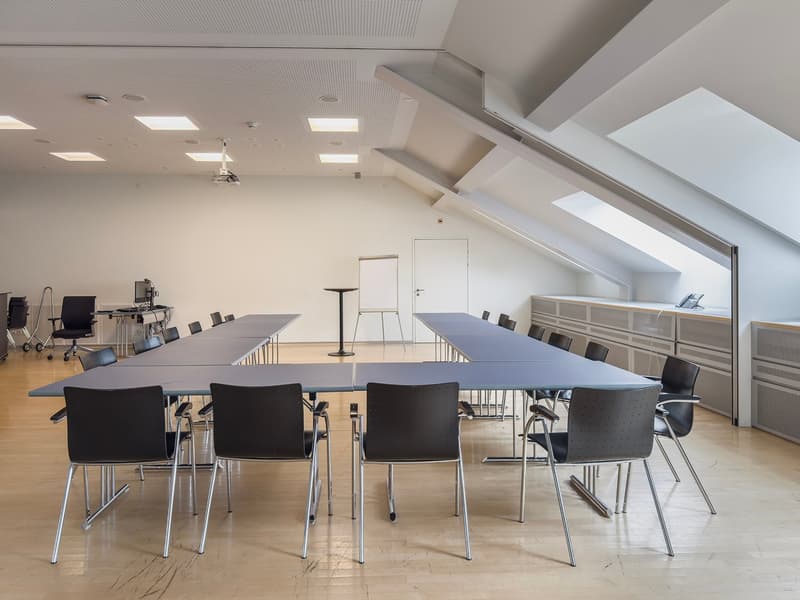 Large open conference room with multiple long tables and black chairs, a whiteboard, and various office equipment and storage units. The room has high ceilings with skylights and a modern, minimalist design.