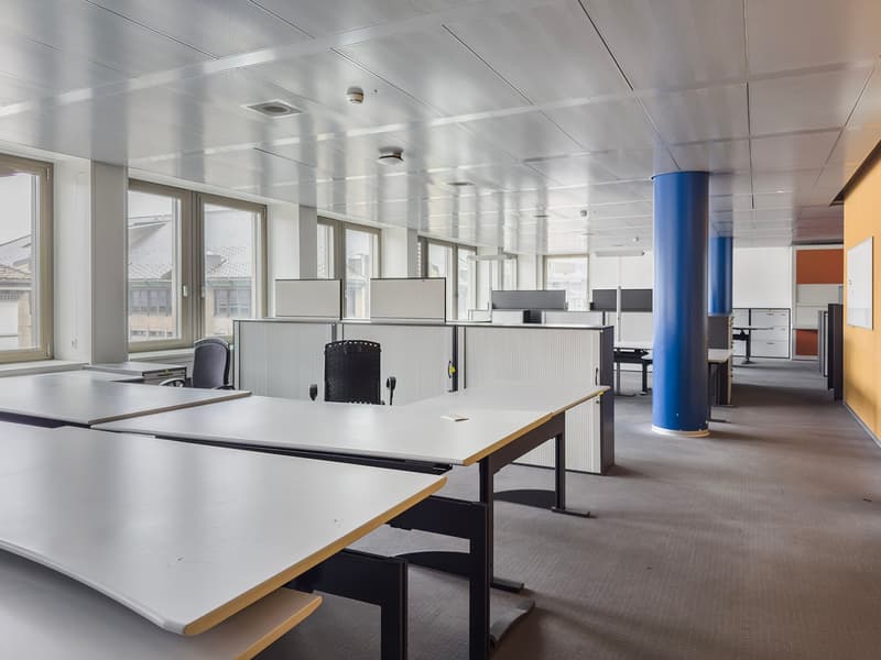 The image shows a modern office space with a large open floor plan. The room has a high ceiling with recessed lighting, and the walls are lined with large windows providing natural light. There are several desks and workstations set up throughout the spac