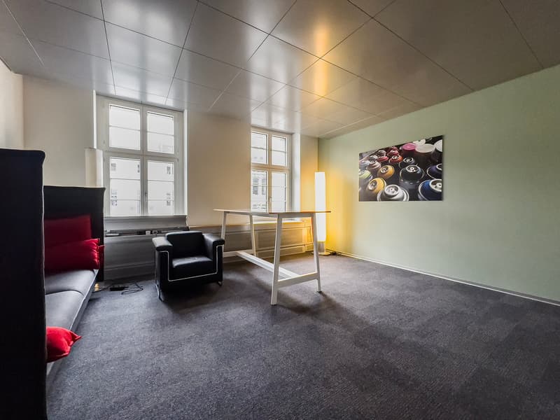 The image shows an office space with a modern and minimalist design. The room has large windows, a gray carpeted floor, and a black leather armchair. There is a wooden desk with a laptop and a piece of artwork on the wall. The ceiling has a grid-like patt