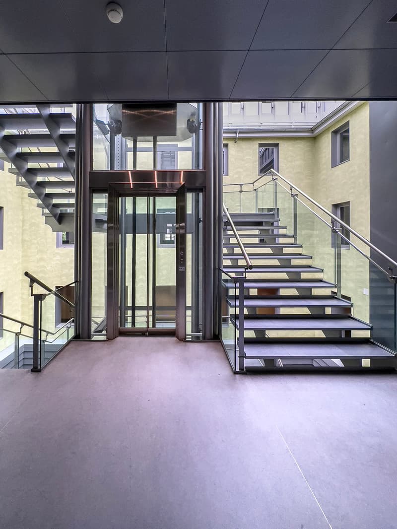 The image shows the interior of a modern building with a glass elevator and a metal staircase. The walls are painted in a light color, and the floor is made of concrete. The space appears to be a lobby or entrance area of the building.