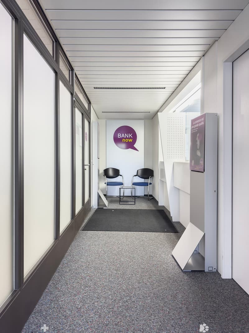 The image shows a hallway or corridor in an office space. The hallway has a gray tiled floor, white walls, and a ceiling with parallel beams. There are two black chairs placed in the hallway, and a large sign on the wall that says 'BANK now'. The hallway