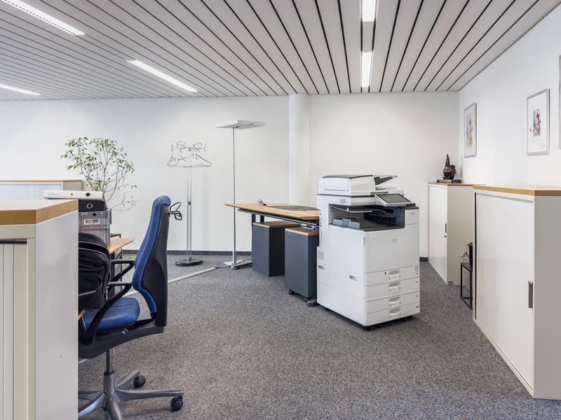 The image shows an office space with a desk, computer, printer/copier, and other office equipment. The ceiling has a distinctive pattern of parallel lines. The walls are white, and the floor is covered in gray carpeting.