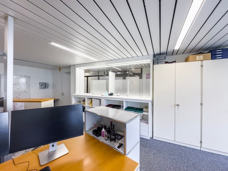 The image shows an office space with a computer monitor, shelves, and storage cabinets. The ceiling has a gray paneled design, and the floor is covered in gray carpeting. The overall space appears to be well-organized and functional for an office environm