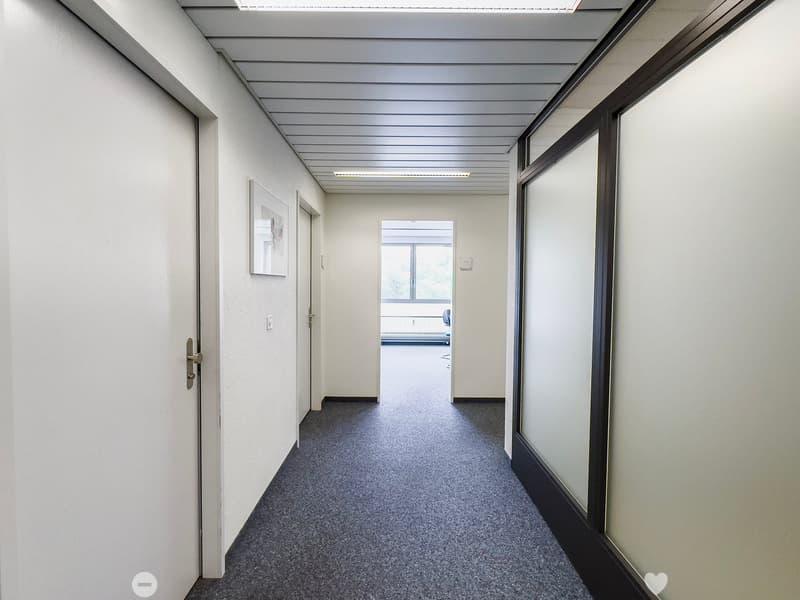 The image shows a long, narrow hallway with a gray terrazzo floor and white walls. The ceiling has a series of parallel beams running across it, creating a modern, industrial look. There are several doors lining the hallway, suggesting this is an office o