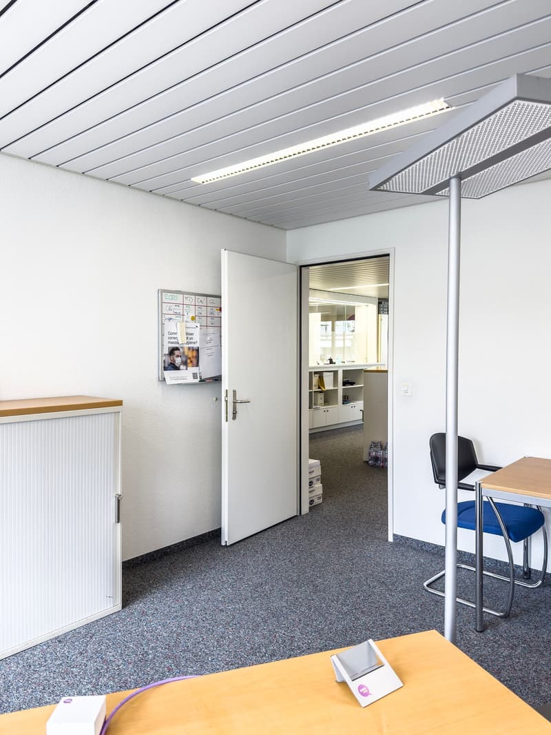 The image shows an office space with a white wall, a gray ceiling with linear lighting fixtures, and a carpeted floor. There is a whiteboard on the wall, a desk with a computer monitor, and a chair. The door to an adjacent room is visible, suggesting this