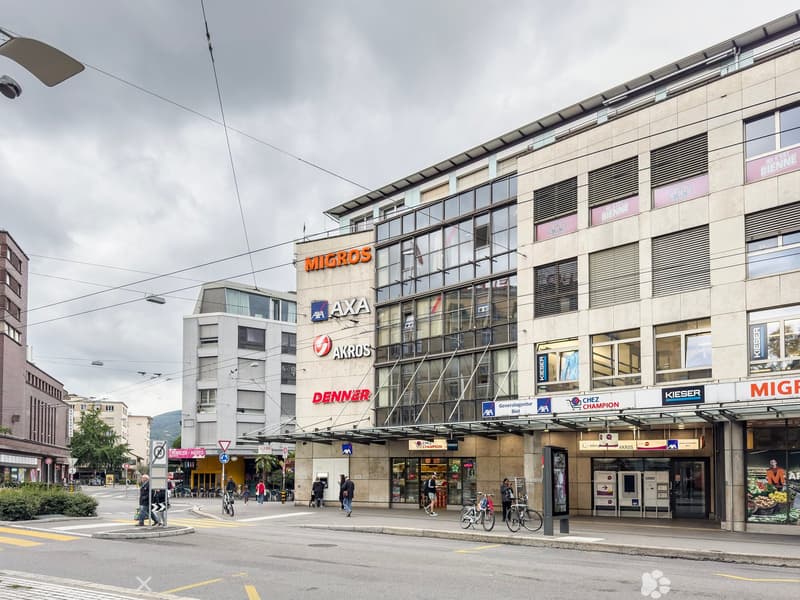Multi-story commercial building with various retail stores and businesses, including Migros, Axa, Akros, and Denner. The building has a modern glass and concrete facade with signage for the different companies. There are people walking and cycling in the