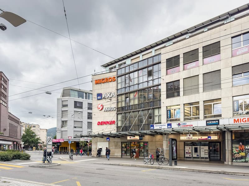 Multi-story commercial building with various retail stores and businesses, including Migros, Axa, Akros, and Denner. The building has a modern glass and concrete facade with signage for the different companies. There are people walking and cycling in the