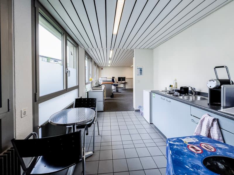 The image shows an office space with a kitchenette area. The space has a tiled floor, white walls, and a ceiling with parallel slats. There is a round table with chairs, a kitchenette with a sink and appliances, and various office equipment and supplies v
