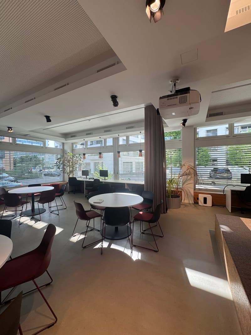 Open office space with large windows, round tables, chairs, and a projector mounted on the ceiling