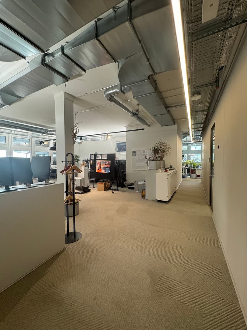 Open office layout with exposed ceiling, carpeted floor, desks, computers, and storage units