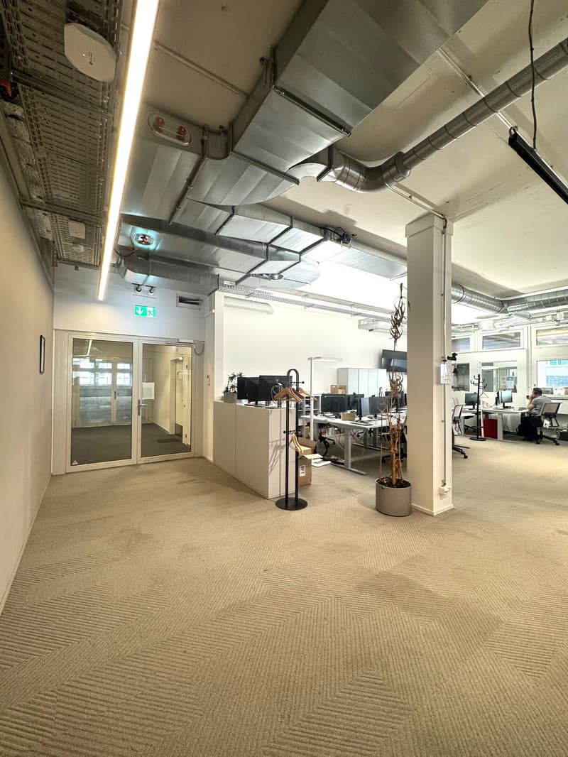 The image shows an open office space with a modern, industrial design. The space features exposed ceiling ducts and pipes, as well as a carpeted floor with a geometric pattern. There are desks and computer equipment visible, indicating this is a workspace