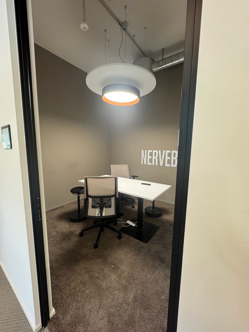 The image shows a small office space with a white desk, a black office chair, and a large 'NERVEB' sign on the wall. The room has a modern, minimalist design with a pendant light fixture and gray walls.