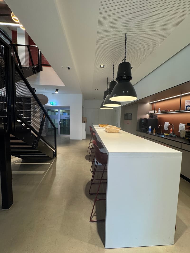 The image shows a modern, open-plan kitchen and dining area. The space features a long, white counter with bar stools, pendant lighting fixtures, and a kitchen area with appliances. The room has a minimalist, industrial-style design with concrete floors a