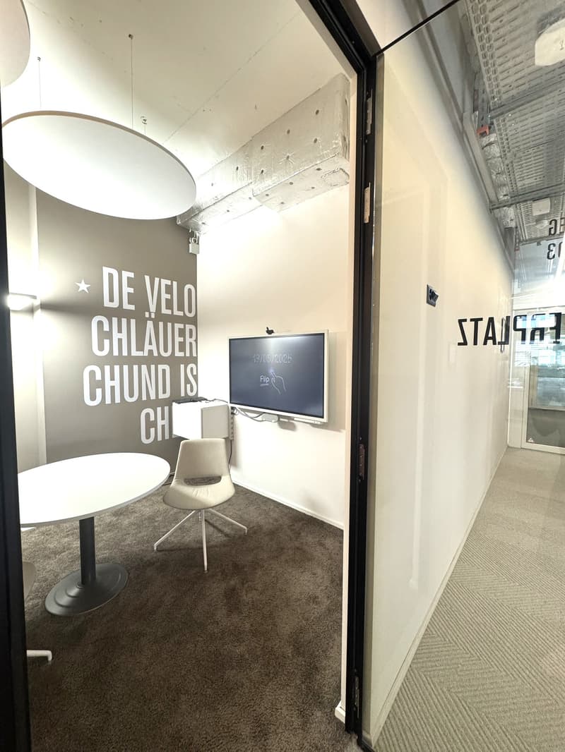 The image shows an office space with a large wall text that says 'DE VELO CHLAUER GHUND IS CH', a TV screen, a white swivel chair, and a carpeted floor.