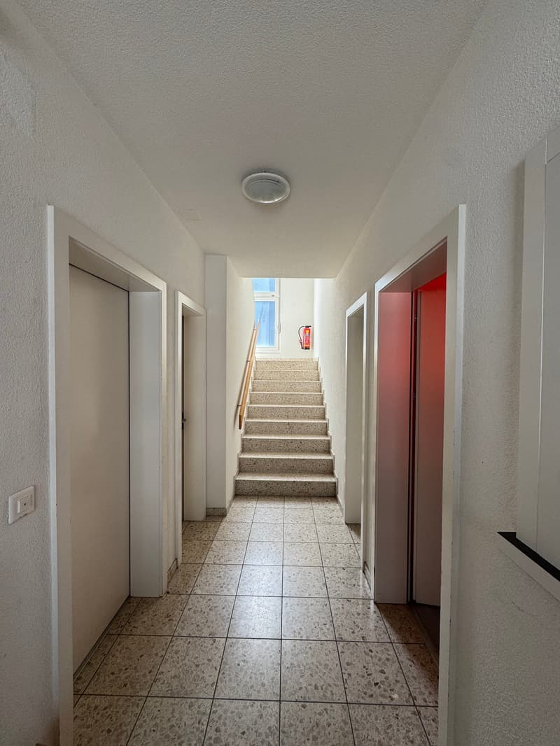 Corridor with tiled flooring, white walls, doorways, a staircase leading to the upper floor, a light fixture on the ceiling, and a fire extinguisher.