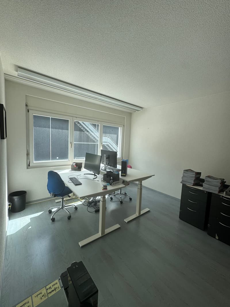An office room with gray walls, white ceiling, two tables, two chairs, two computer monitors, and black cabinets
