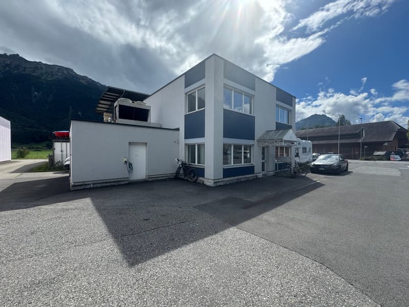 A two-story building with a white exterior and blue accents. There is a garage on the left side of the building with a white door. To the right of the building, there is a covered parking area with a car parked in it. A bicycle is parked in front of the b