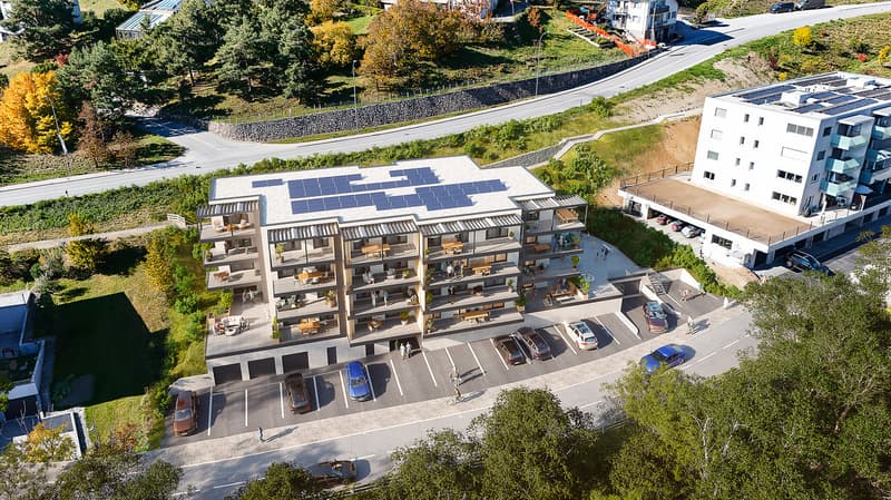 Aerial view of a multi-story building with solar panels on the roof, multiple balconies, and a parking lot with several cars parked.