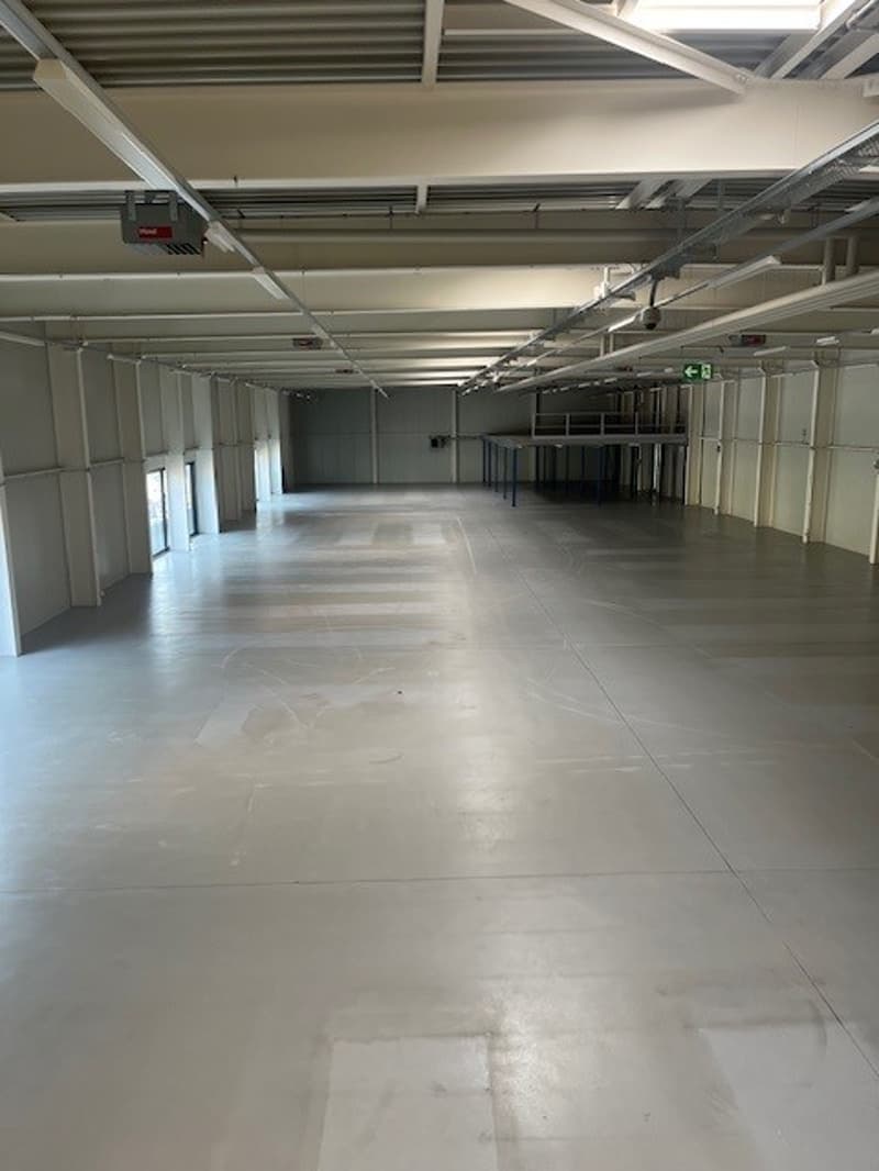 Large open warehouse space with concrete flooring, exposed ceiling beams, and overhead lighting. The space appears to be empty and ready for use.