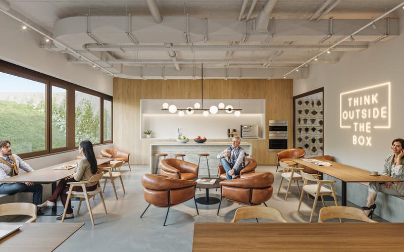Open concept office space with exposed concrete ceiling, wooden walls, and large windows. Furnished with comfortable leather sofas and chairs, a kitchen area with a dishwasher, and a neon sign that says 'Think outside the box'. Several people are working