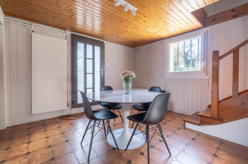 Dining area with a round white table and black chairs. Wooden walls and ceiling. Wooden staircase to upper level. Windows with white frames.